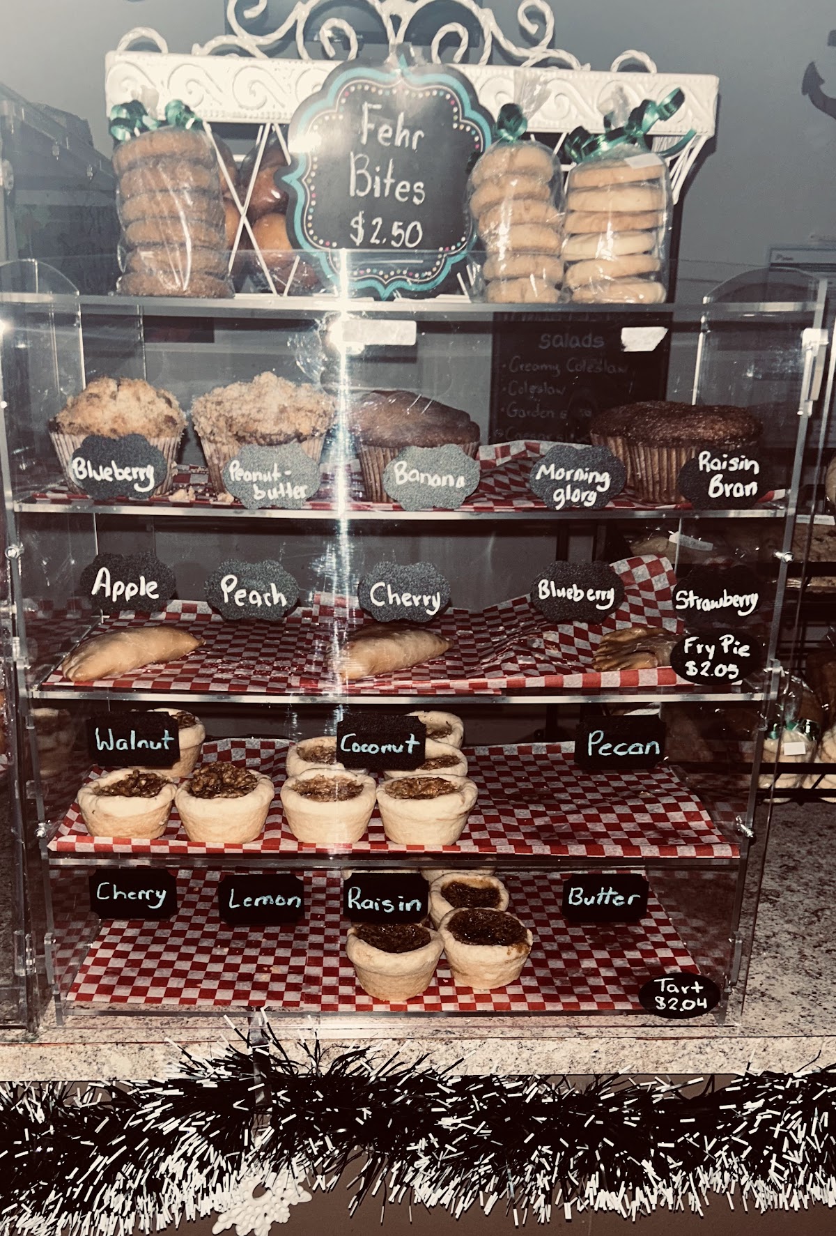 Full display case at Fehr's Heritage Bakery with Fehr Bites muffins, fruit pies on checkered paper, and rows of butter tarts with chalkboard labels