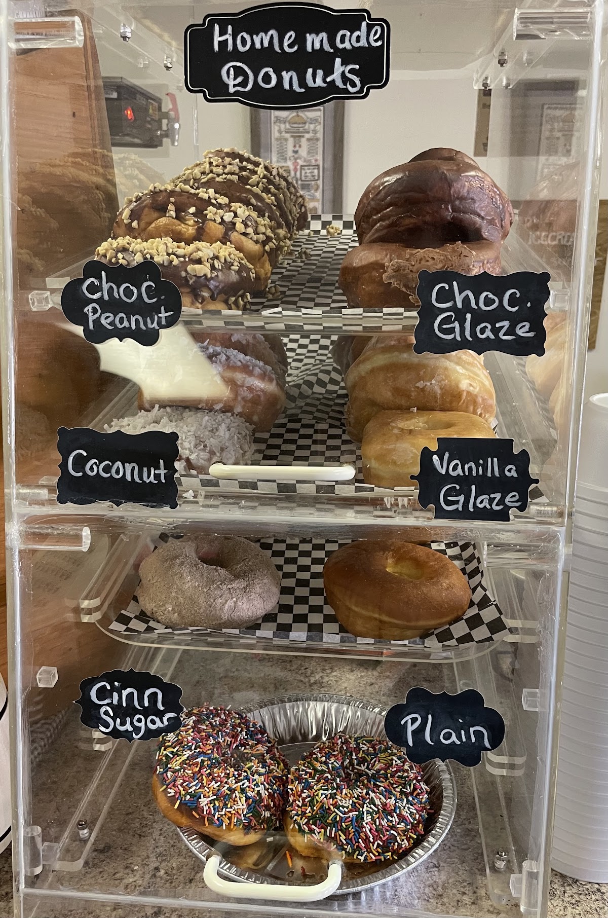 Homemade donuts display at Fehr's Heritage Bakery with chalkboard labels showing Choc Peanut, Choc Glaze, Coconut, Vanilla Glaze, Cinn Sugar, and Plain varieties